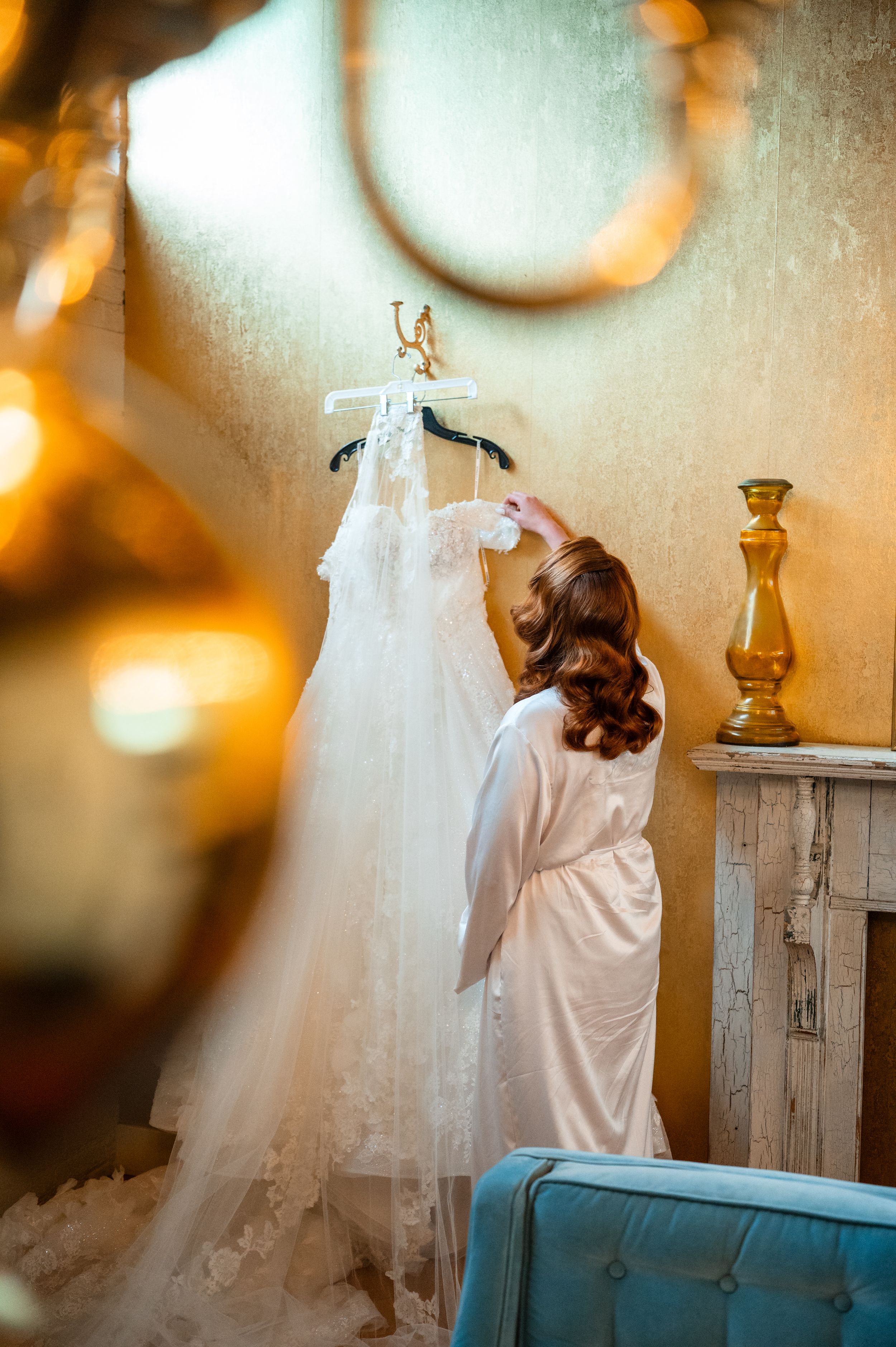Bride in white robe reaches for lace wedding dress hanging on wall in golden lit room.