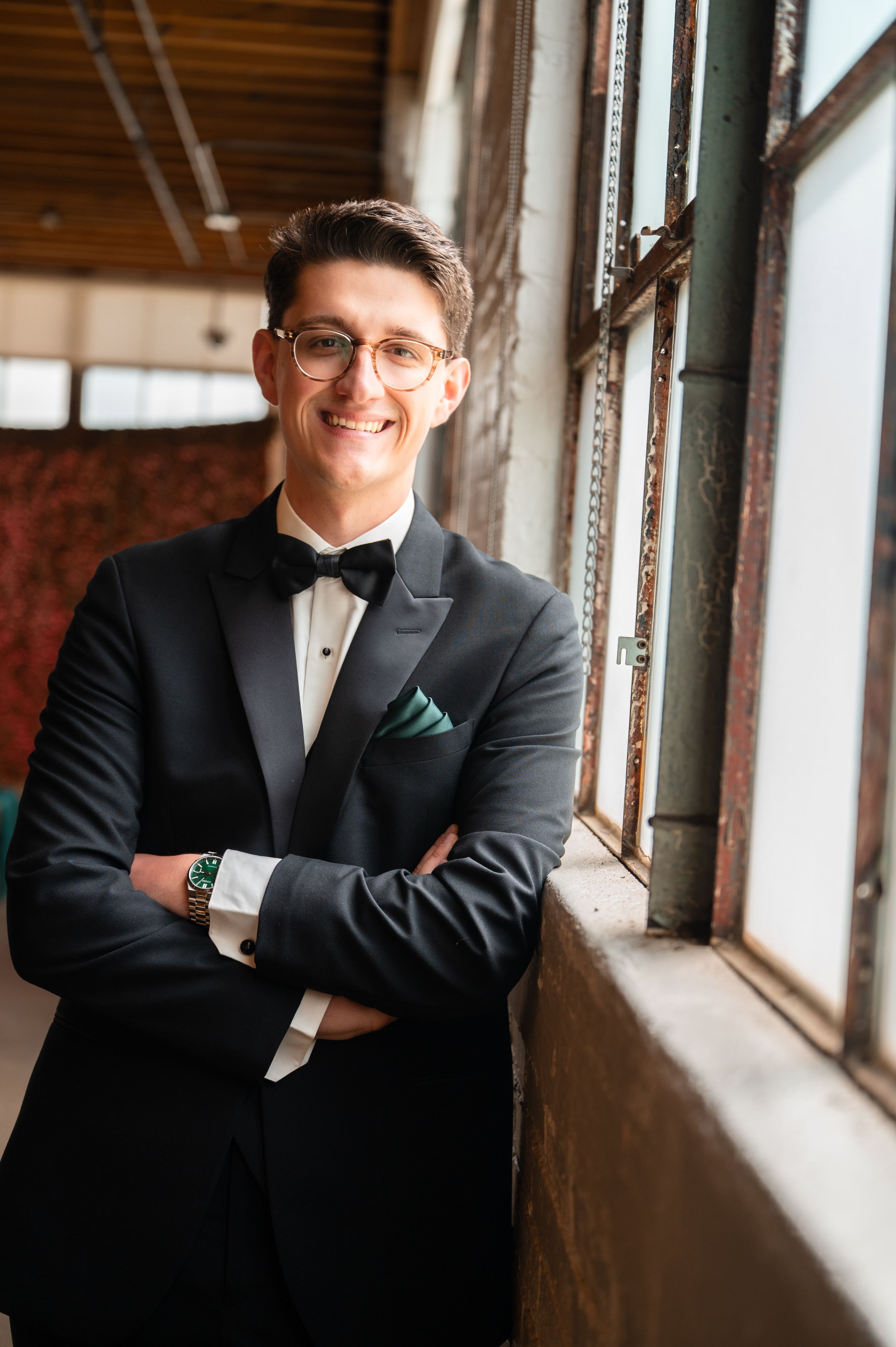 Dapper groom in black tuxedo with bow tie and green pocket square poses by industrial window.