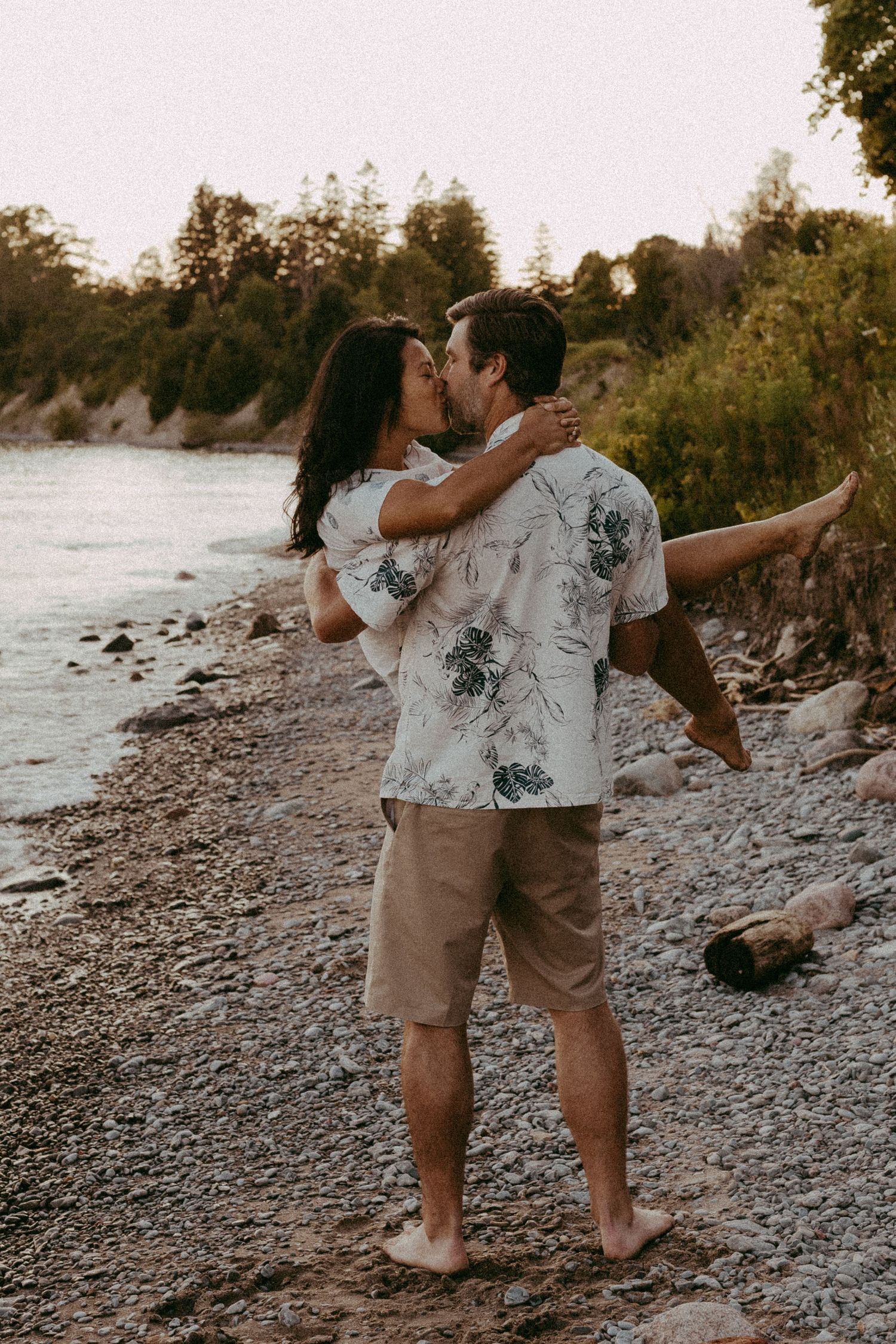 A couple shares a romantic kiss on a rocky beach shoreline, man in floral shirt holding woman in his arms.