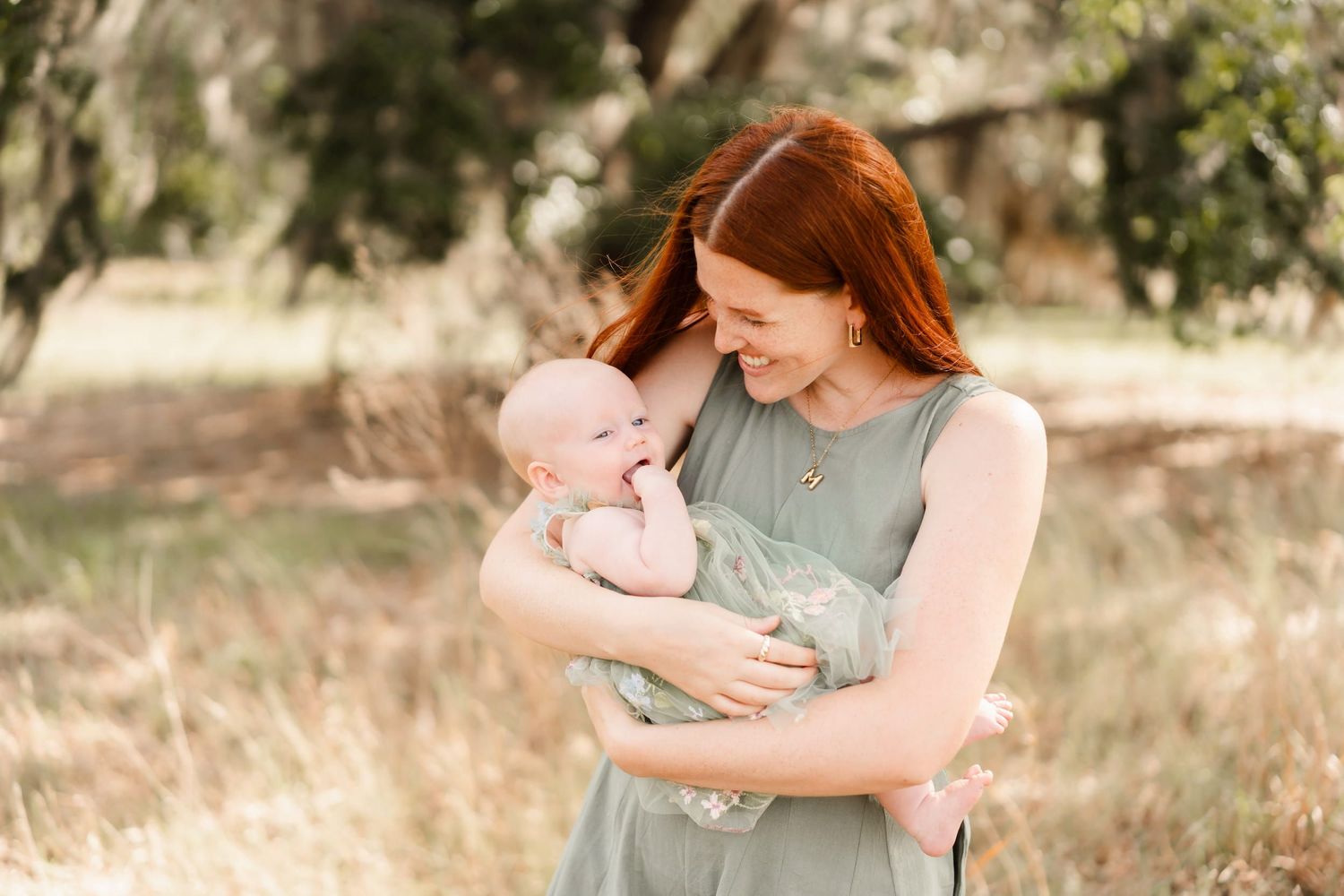 Red-haired mother smiling tenderly at her newborn baby outdoors in a sunlit green field.