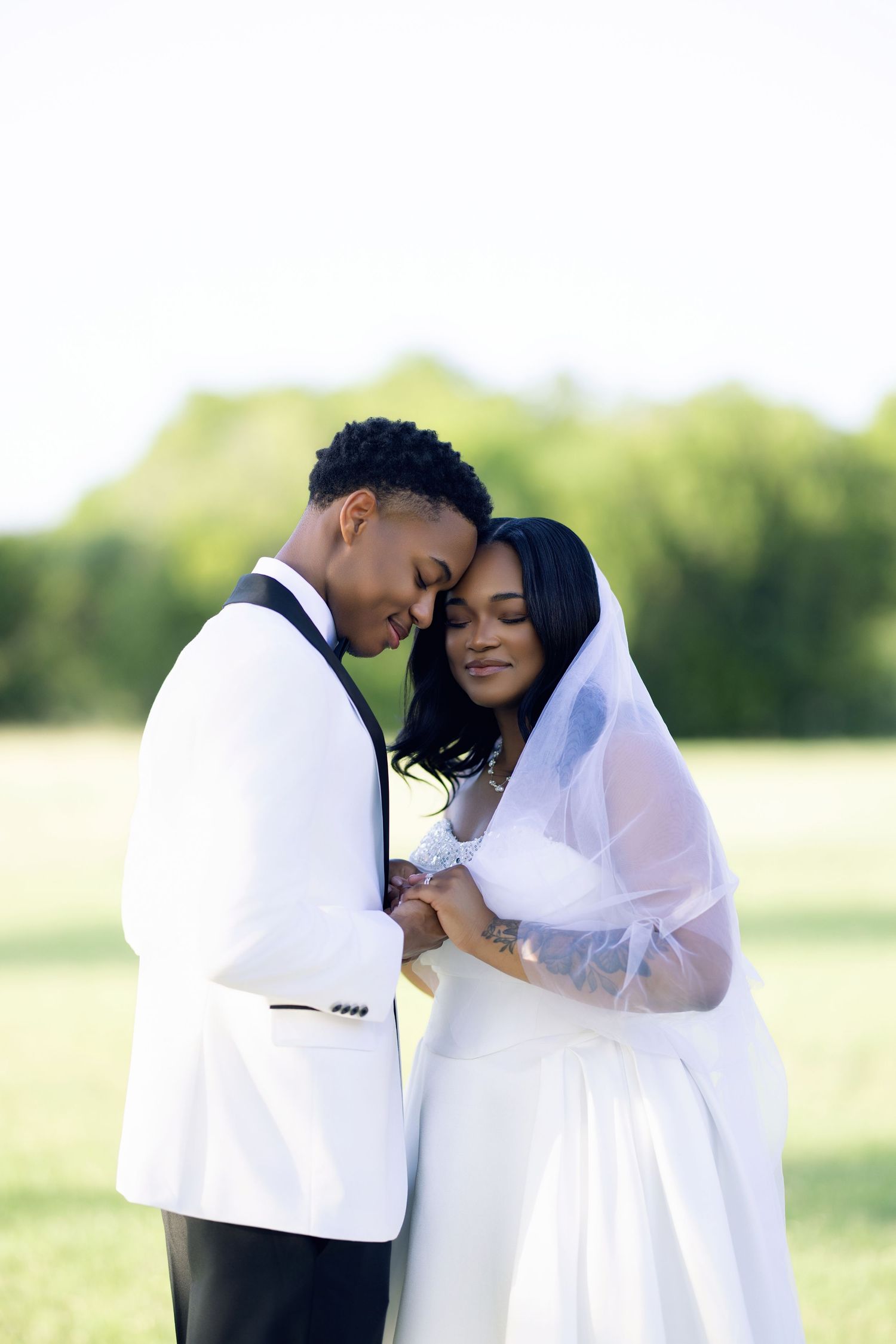 A Black couple shares a tender moment in wedding attire outdoors, bride in white gown and veil, groom in white tuxedo jacket.