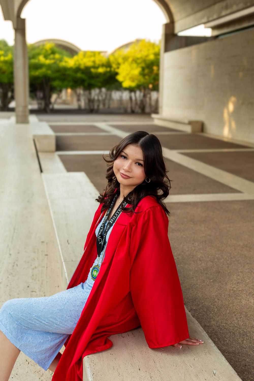 Young woman in red graduation gown sitting on steps outdoors, smiling at camera during graduation photo session.