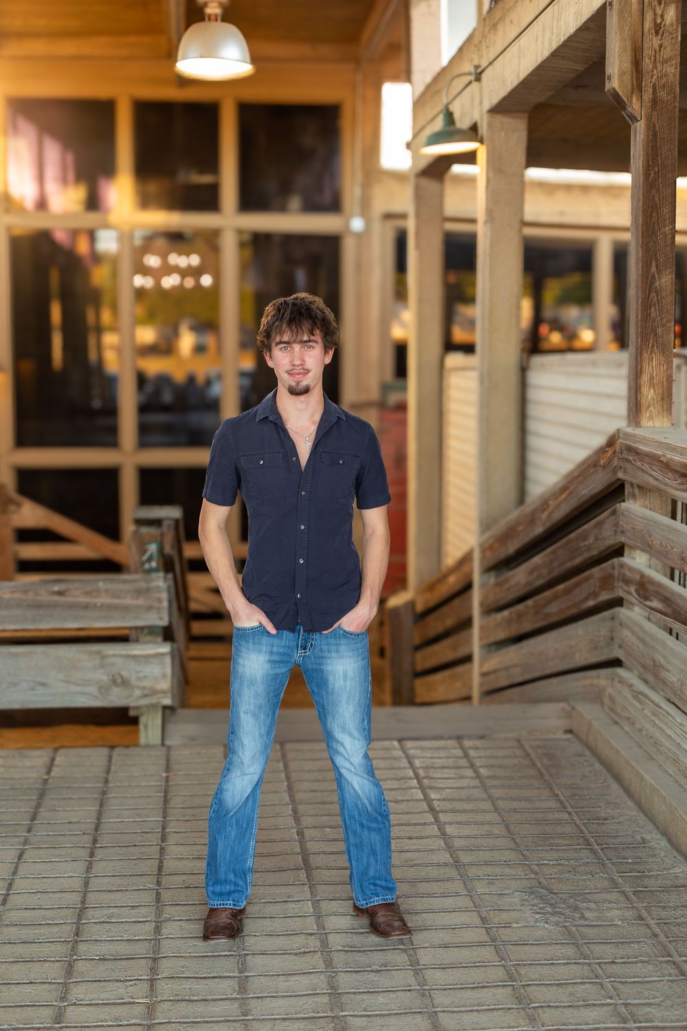 Young man in blue shirt and jeans standing confidently outside a rustic wooden building with hands in pockets.