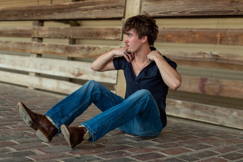 Young man in blue jeans and dark shirt sitting casually on brick floor against wooden wall backdrop.