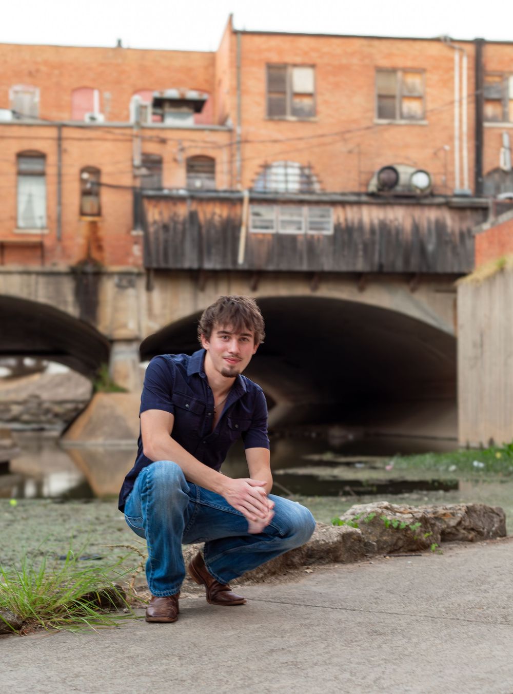 Young man in blue shirt and jeans crouching near a bridge over a creek in an urban setting.