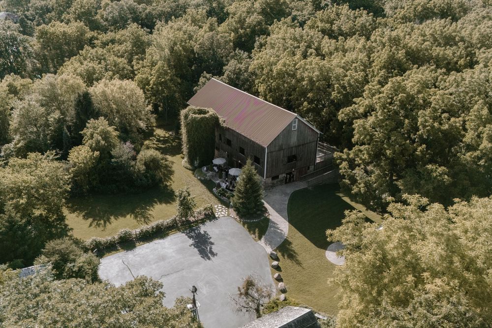 Aerial view of a rustic stone barn venue surrounded by lush green trees with a paved parking area.