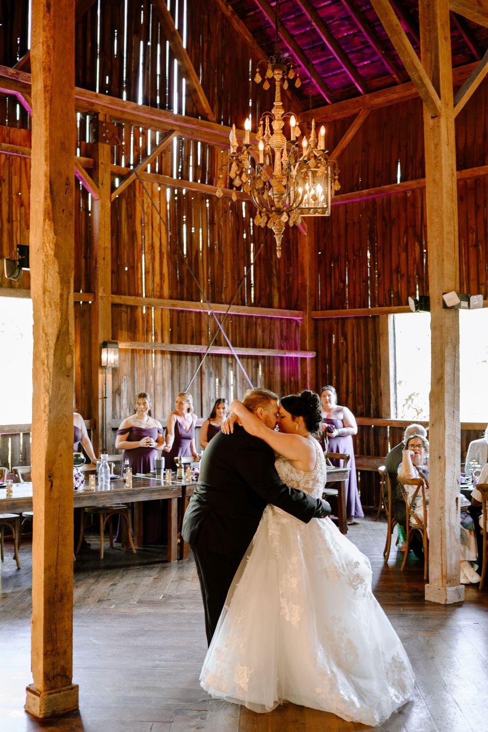 Bride and groom share first dance under chandelier in rustic barn wedding venue with bridesmaids in purple.