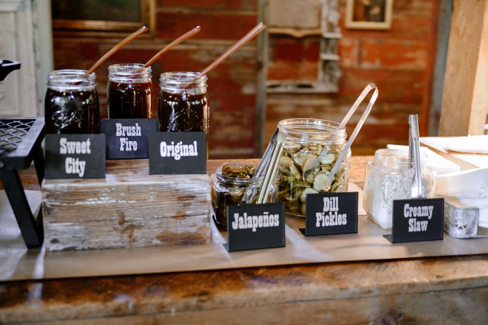 Rustic condiment bar with mason jars of sauces labeled Sweet City, Break Fire, Original, jalapenos, dill pickles, and creamy slaw.