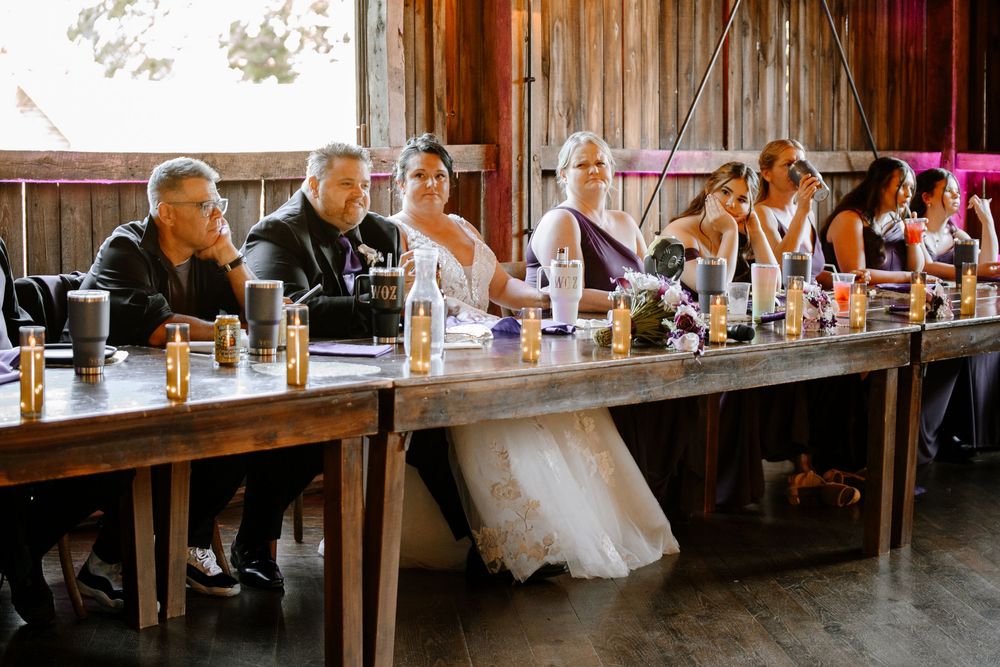 Wedding party seated at rustic wooden head table during barn reception with golden candlelight ambiance.