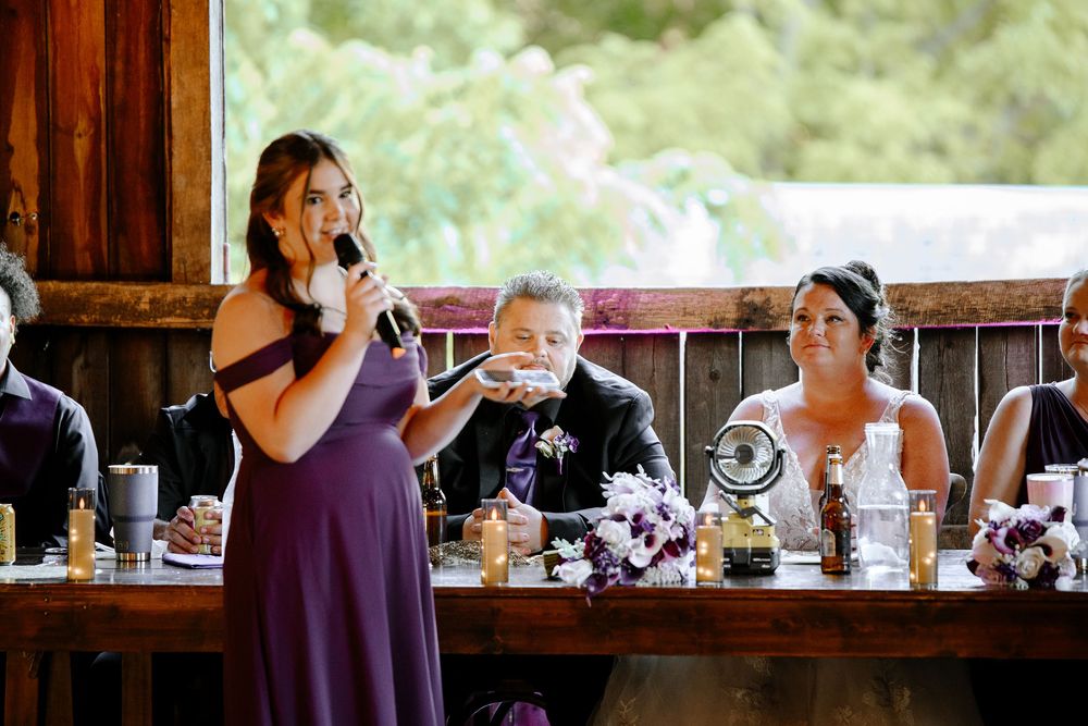 Bridesmaid in purple dress gives wedding toast as bride laughs at decorated head table.