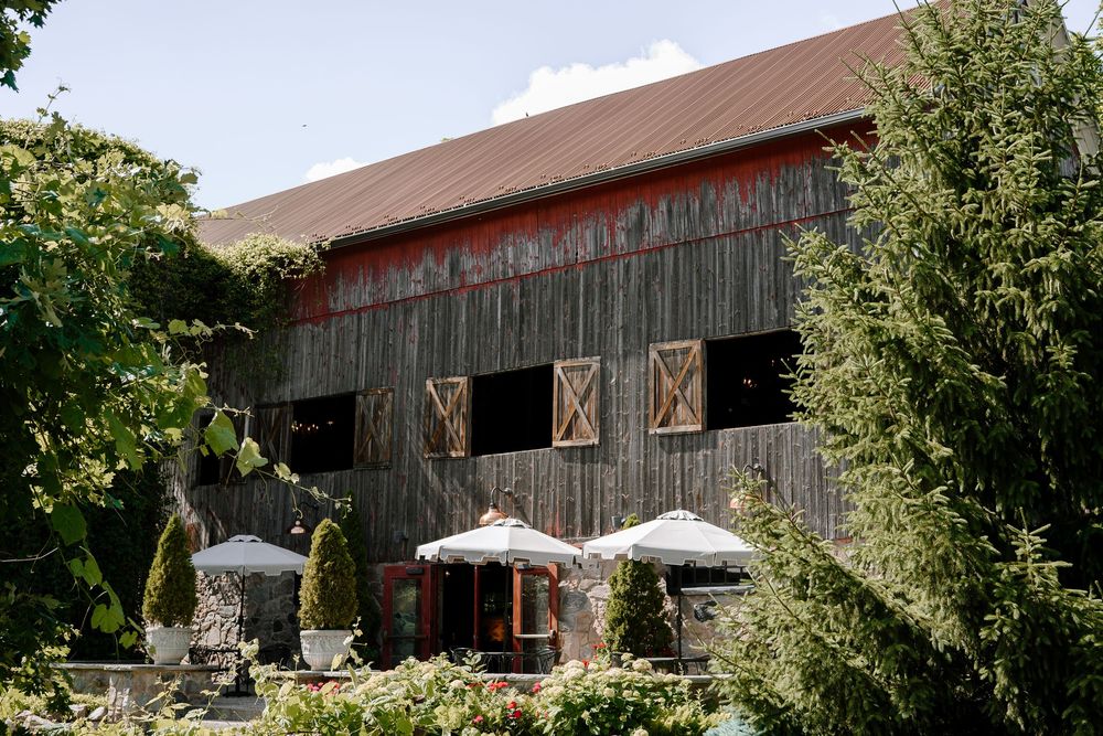 Rustic weathered barn venue with red trim, decorative shutters, and outdoor seating with white umbrellas surrounded by lush greenery.