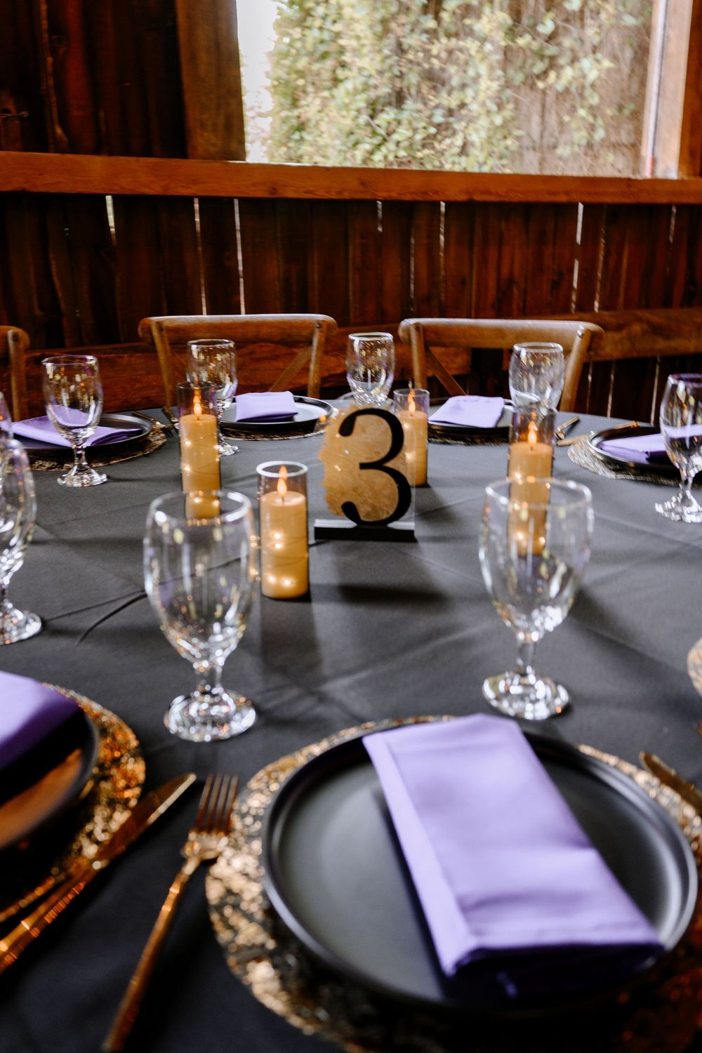 Elegant wedding table setting with purple napkins, gold chargers, candles, and table number 3.