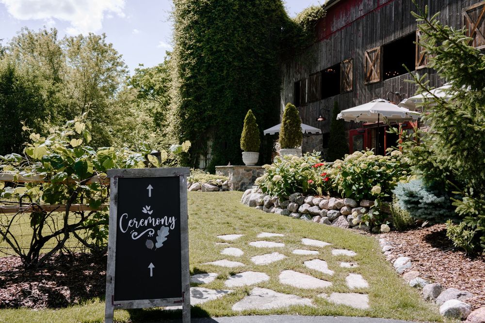 A chalkboard ceremony sign on a stone path leading to a rustic barn venue surrounded by lush greenery.