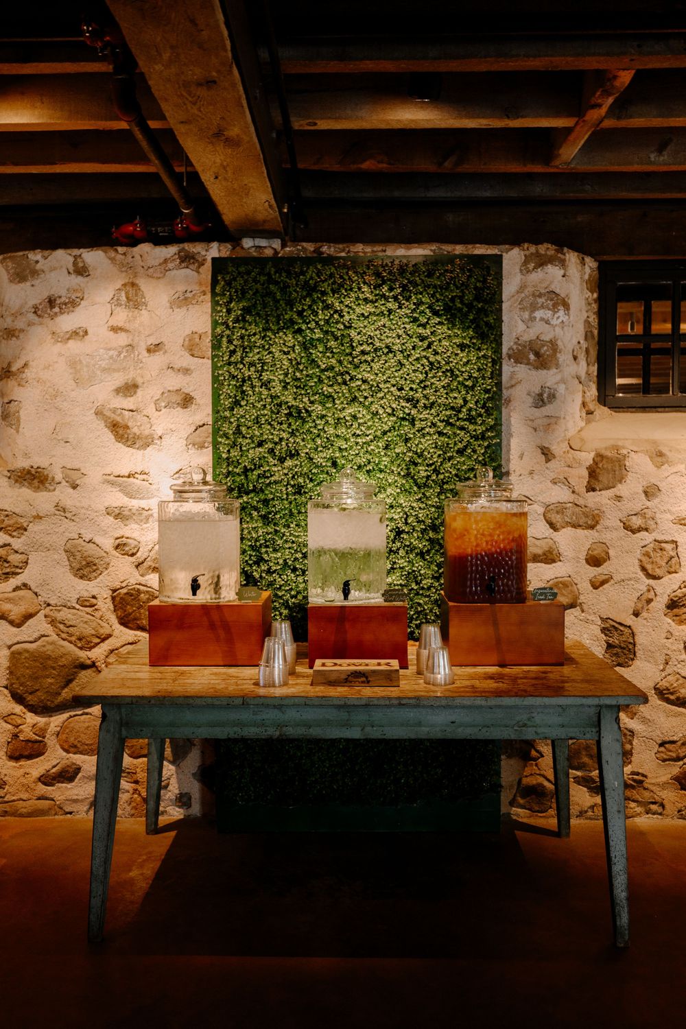 Rustic beverage station with drink dispensers on wooden table against stone wall with greenery backdrop.