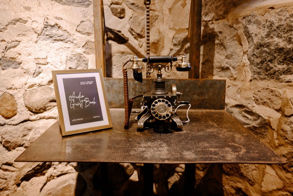 Vintage black rotary telephone beside a framed guest book sign on rustic wooden table against stone wall.