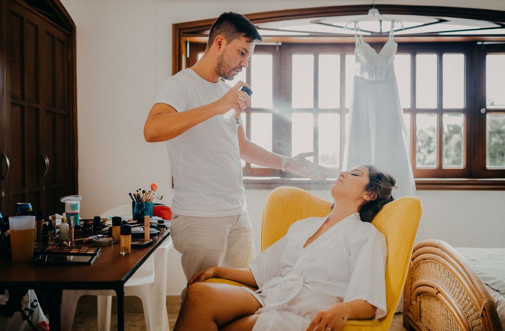 Man applies makeup to a smiling bride in a white robe seated in a yellow chair during wedding preparations.