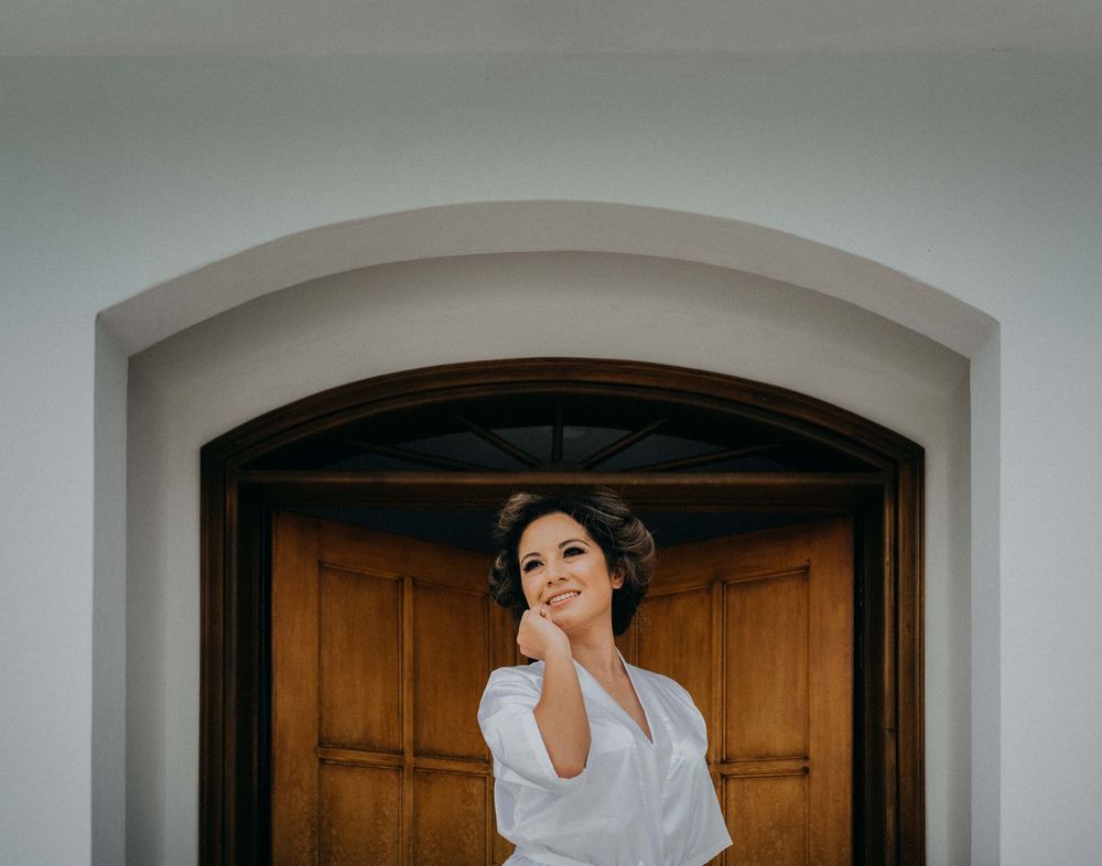 Elegant bride in white robe smiles confidently standing before ornate wooden doors in a bright archway.