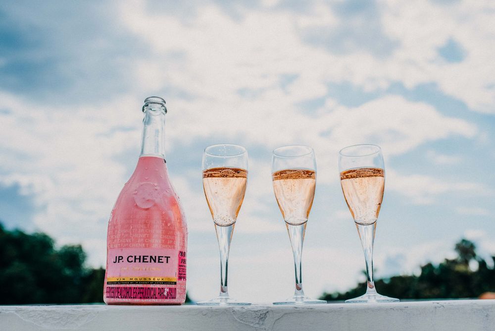 JP. Chenet pink sparkling wine bottle with three filled champagne flutes against a cloudy sky backdrop.