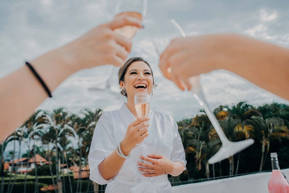 A laughing bride in white holds champagne outdoors, framed by toasting glasses, with palm trees in the background.