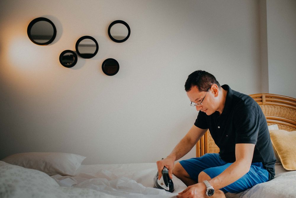 Man in black polo shirt and blue shorts sitting on bed ironing clothes in a bright modern bedroom.