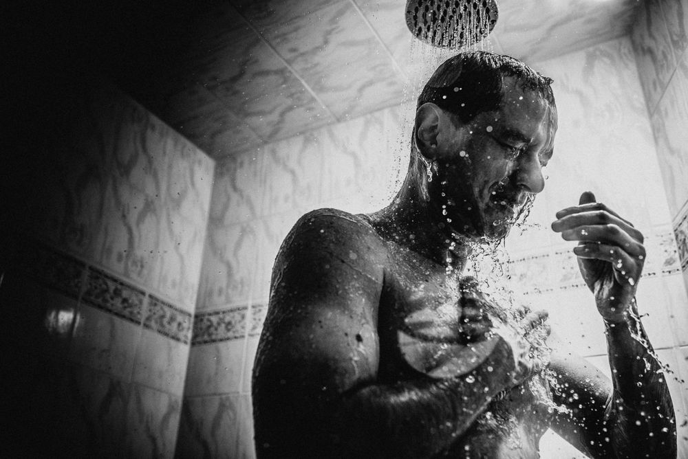 Black and white photo of a man showering under a rain shower head, water cascading down his face.