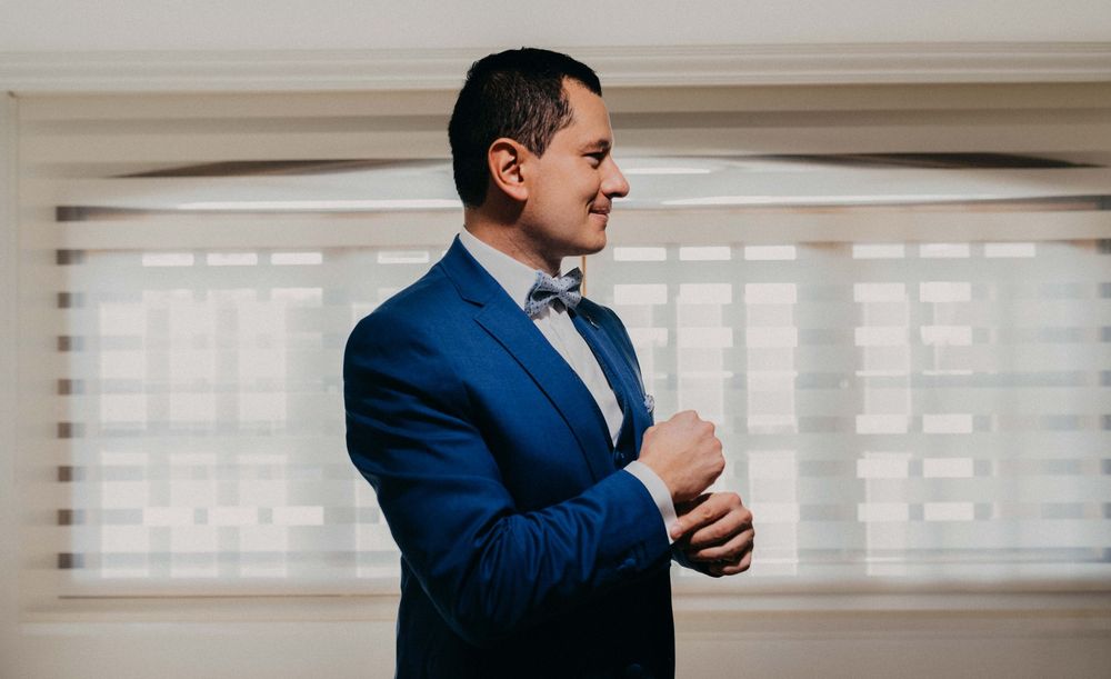 Man in blue suit adjusting cufflinks near window, wearing bow tie, preparing for formal event.