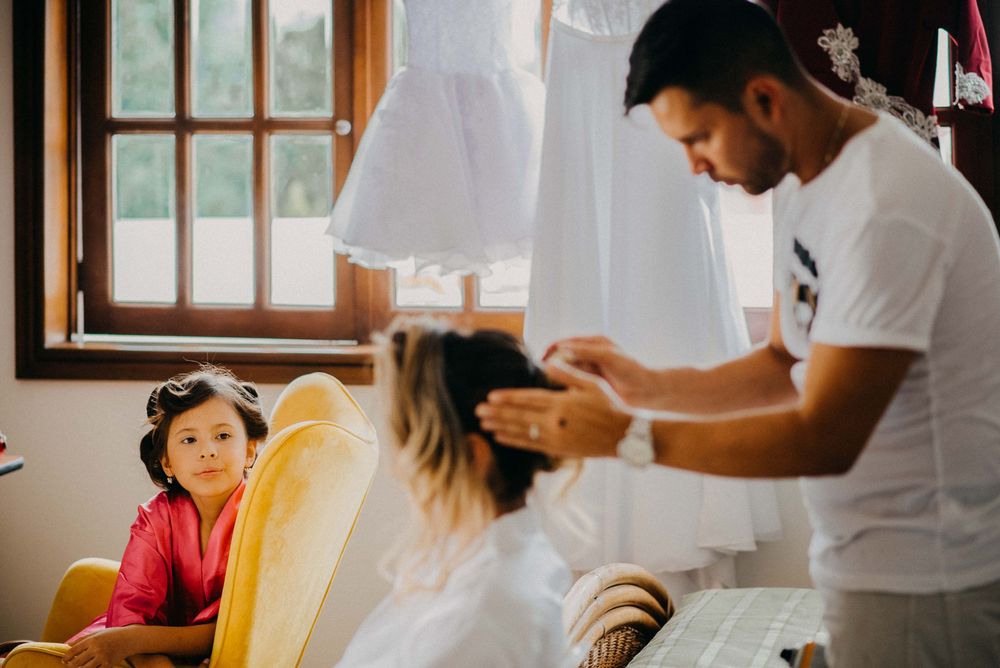 A man styles a woman's hair indoors while a young girl in pink watches from a yellow chair near a window.