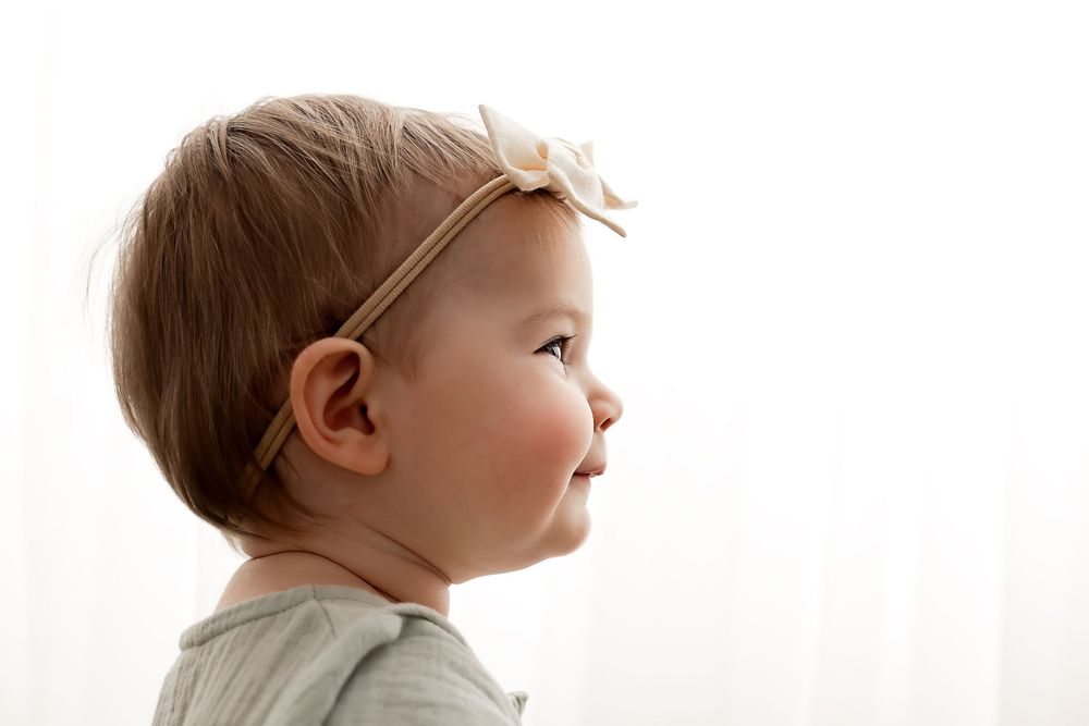 A baby girl in profile wearing a small bow headband, looking upward against a soft white background.