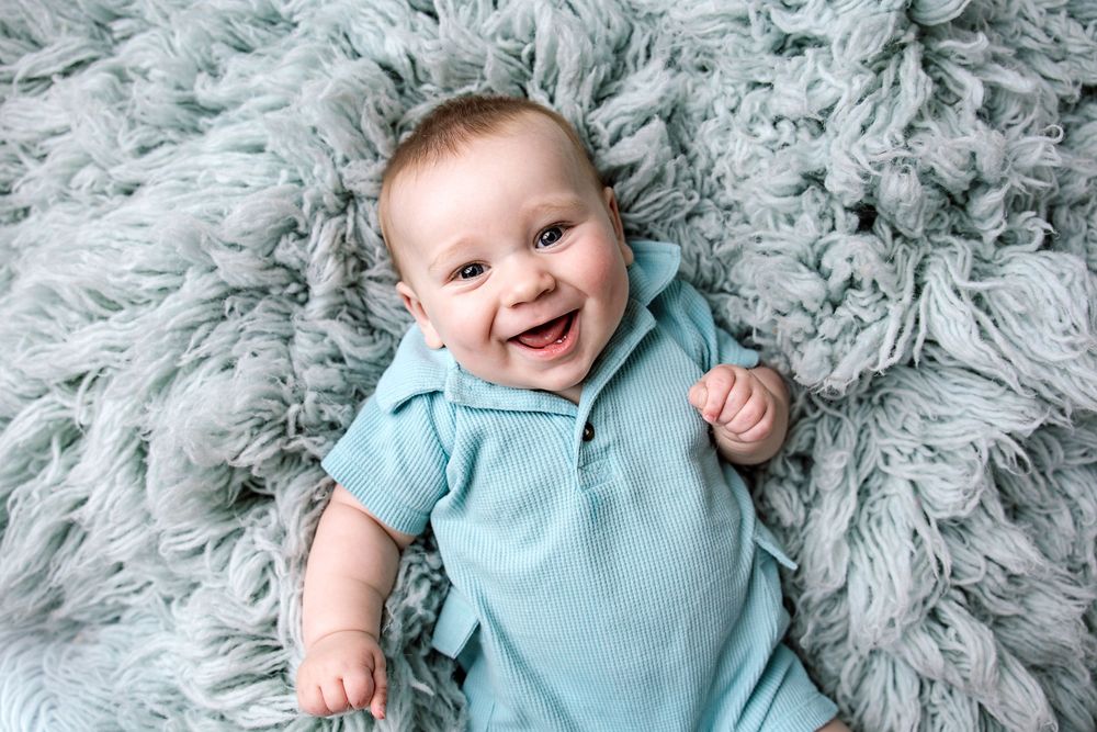Smiling baby in blue onesie lying on fluffy gray fur blanket, looking up with joyful expression.