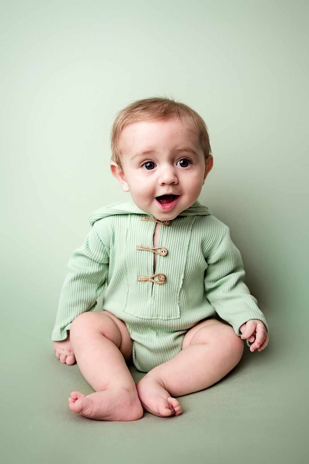 A happy baby in a green ribbed hoodie onesie sits barefoot on a green background, smiling with mouth open.