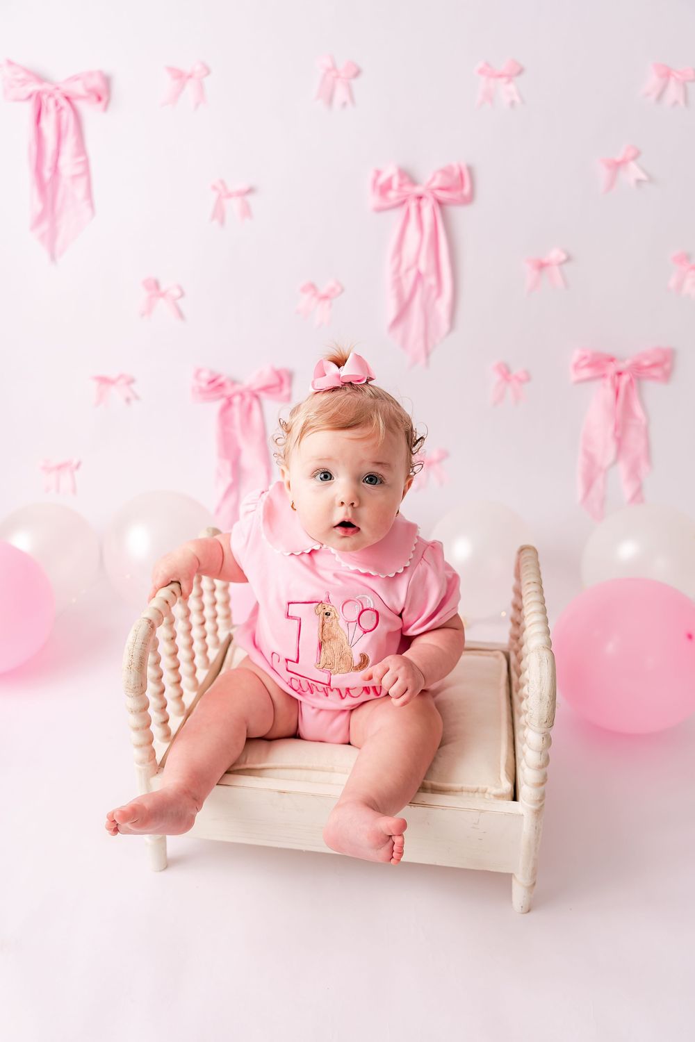 Baby girl in pink onesie sits on white mini bed, surrounded by pink bows and balloons for her 1st birthday.