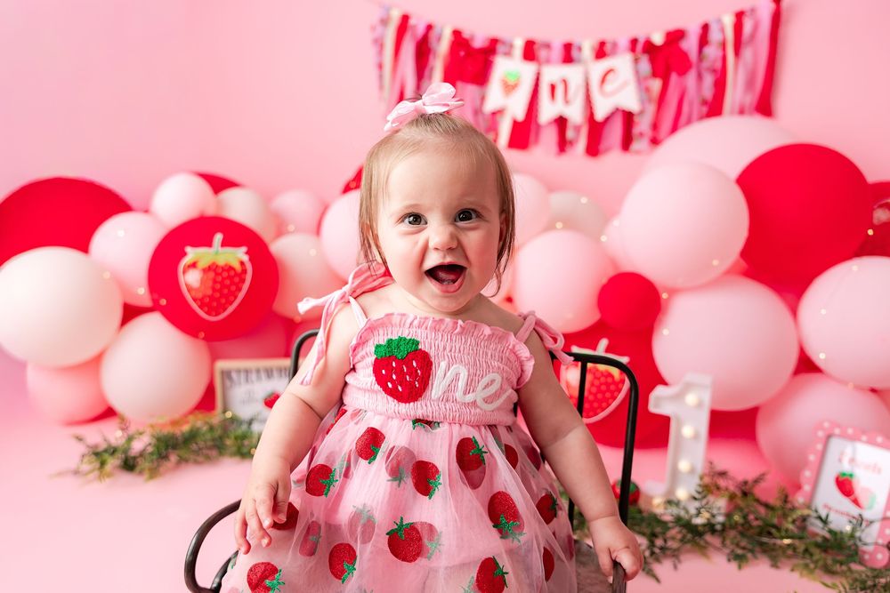 Baby girl in strawberry dress smiles at first birthday cake smash with pink and red balloon backdrop.