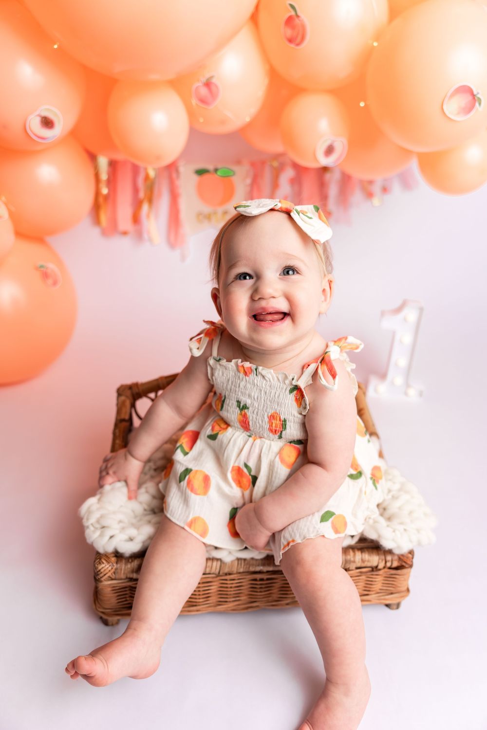 Smiling baby girl in peach fruit dress sits in wicker basket at first birthday party with orange balloons.