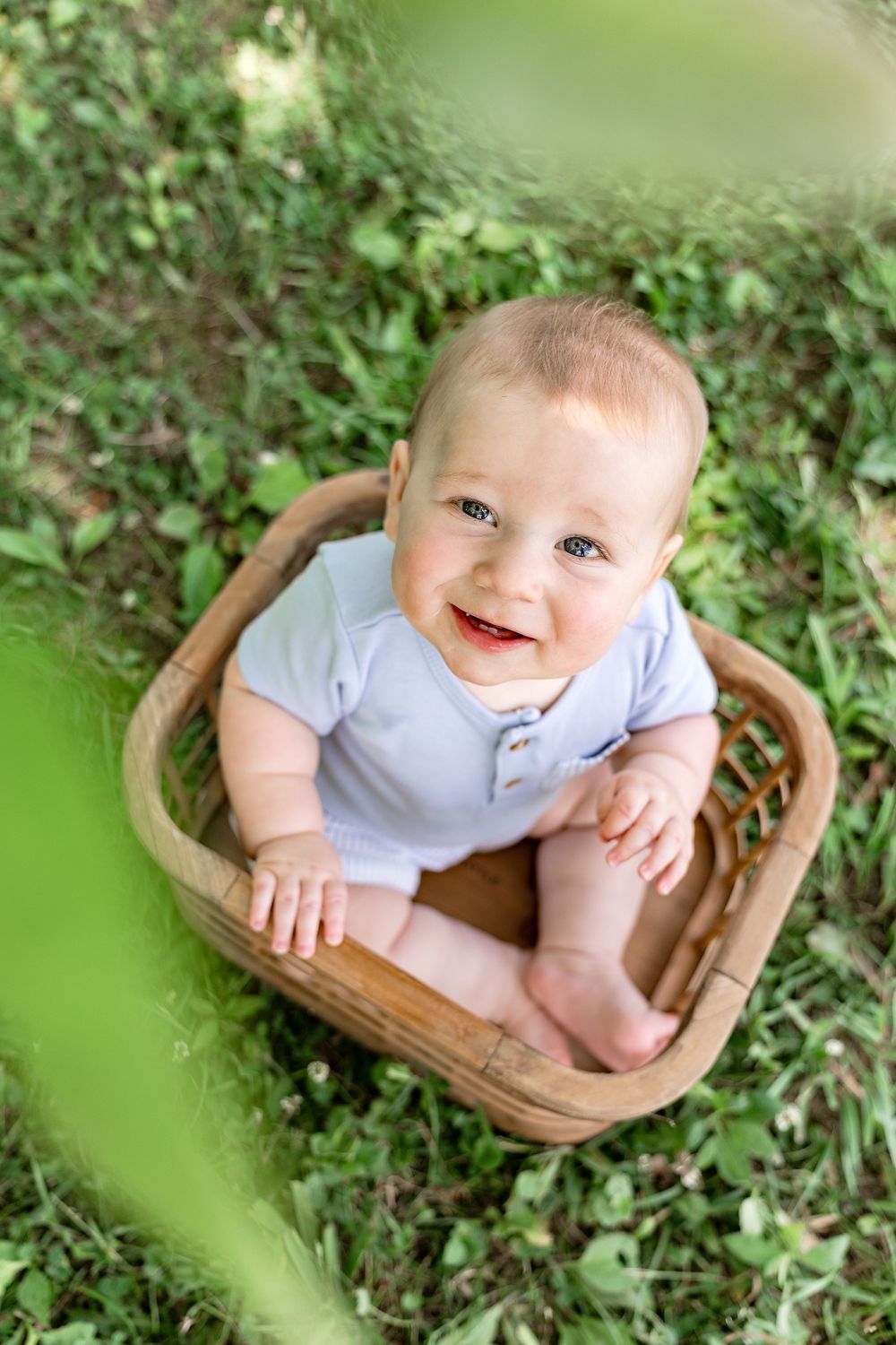 Smiling baby in blue onesie sitting in a rustic wooden basket on green grass outdoors.