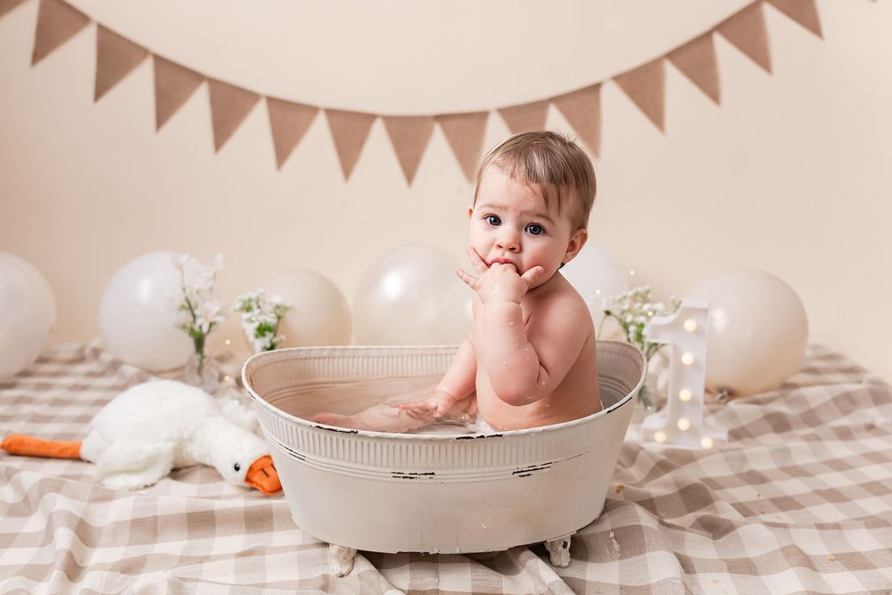 Baby sitting in a vintage wash basin during a first birthday cake smash photo session with balloons and bunting.