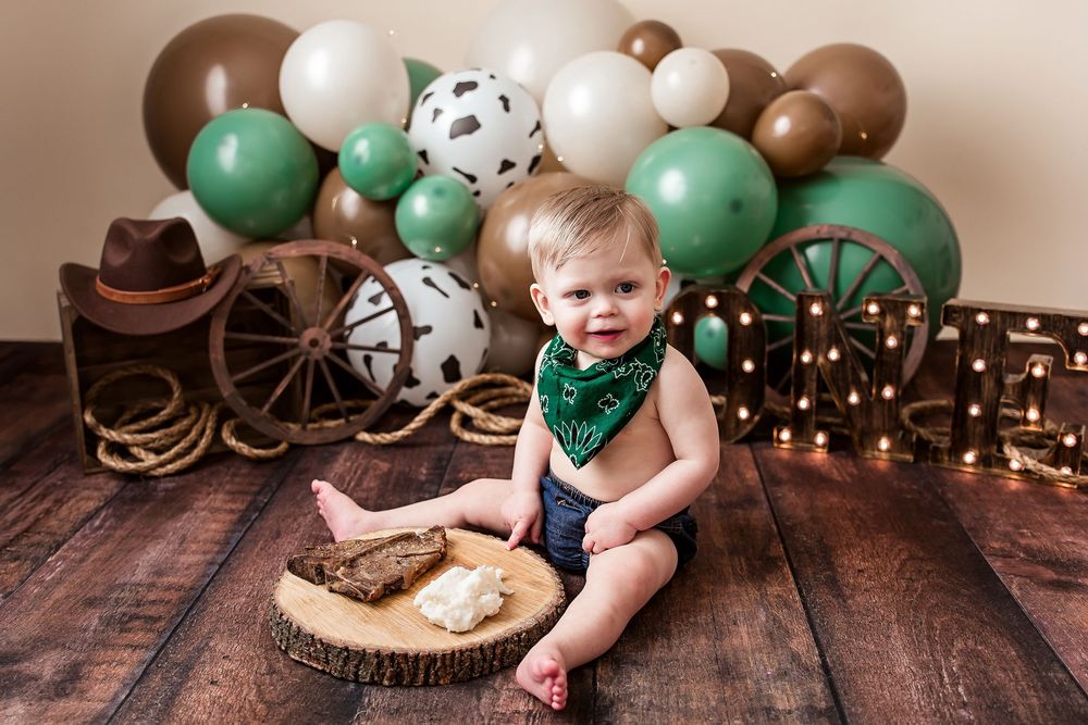 Baby boy in green bandana sits on wood slice with smash cake, surrounded by western-themed balloons and cowboy decor.