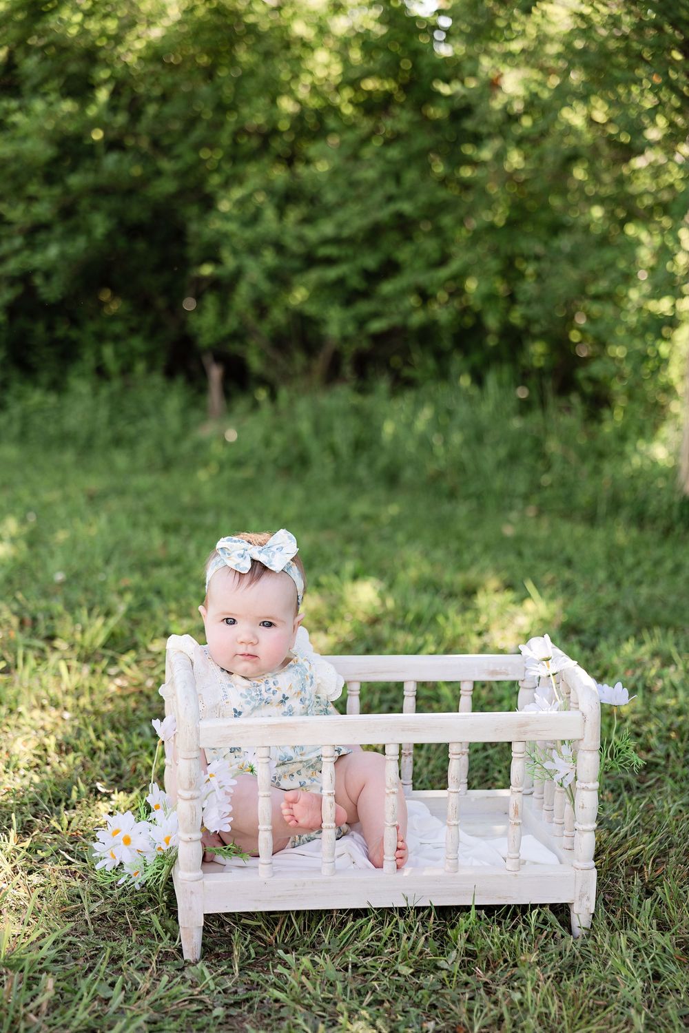 Adorable baby girl in floral outfit and bow headband sitting in a miniature white wooden crib on green grass outdoors.