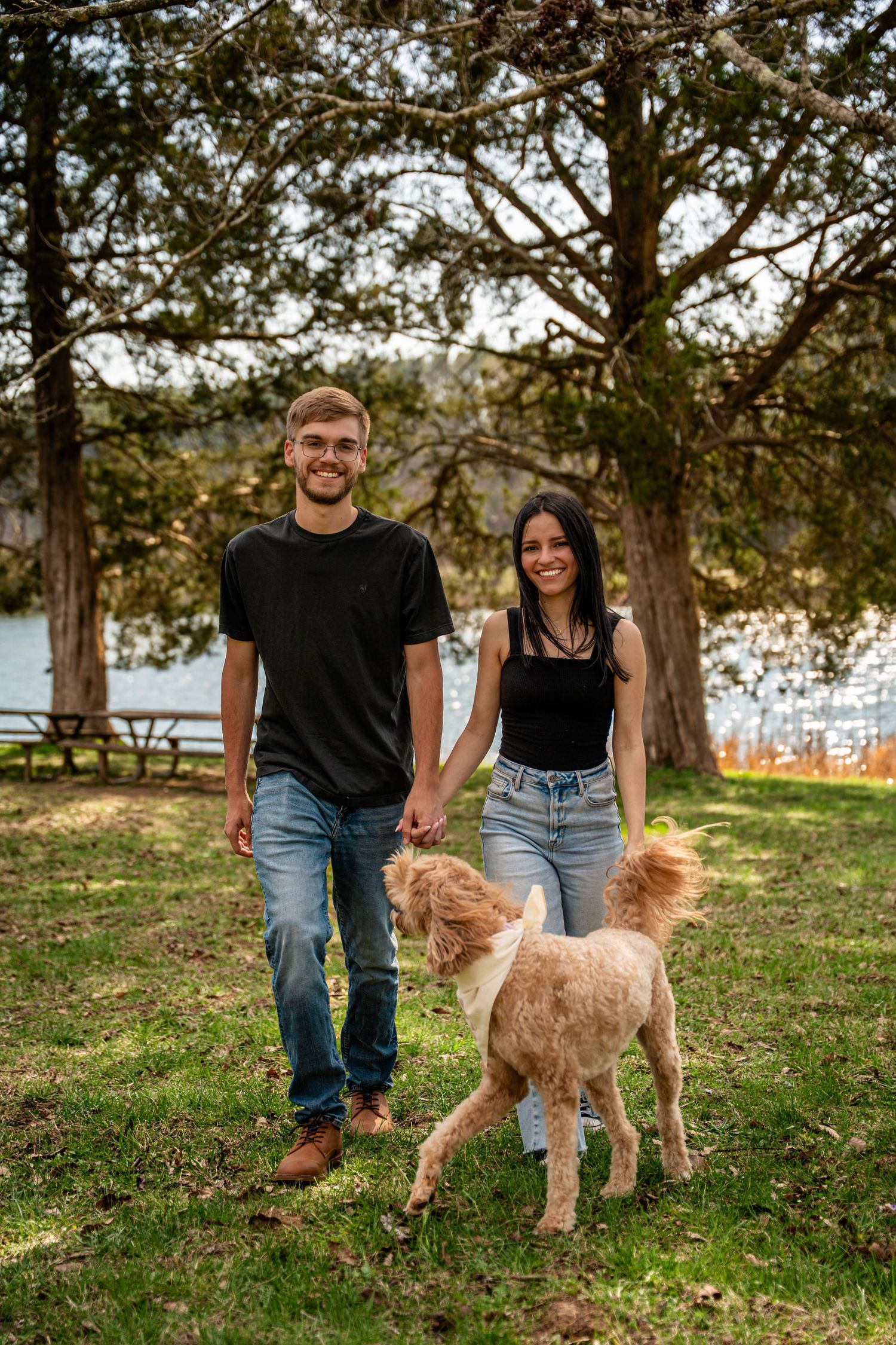 Smiling couple holds hands while walking fluffy dog in scenic lakeside park setting.