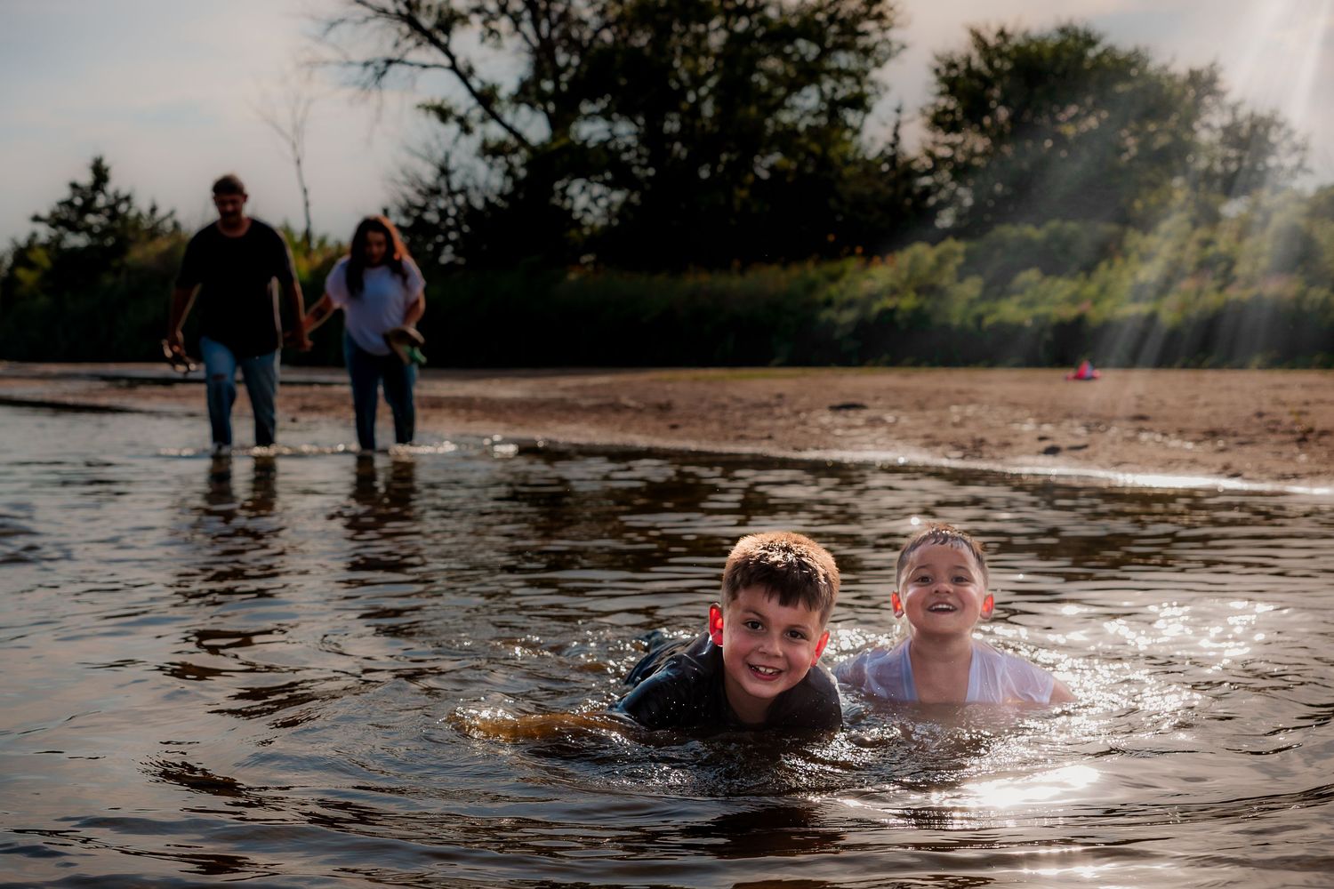 Two children swimming in shallow water at a beach while adults walk behind them in golden sunlight.
