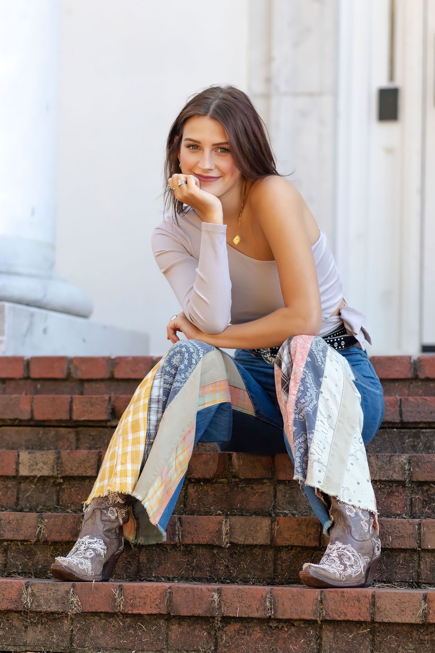 Senior photographer Rome GA Young woman in jeans and cowboy boots sitting on brick steps, smiling outdoors near white columns.