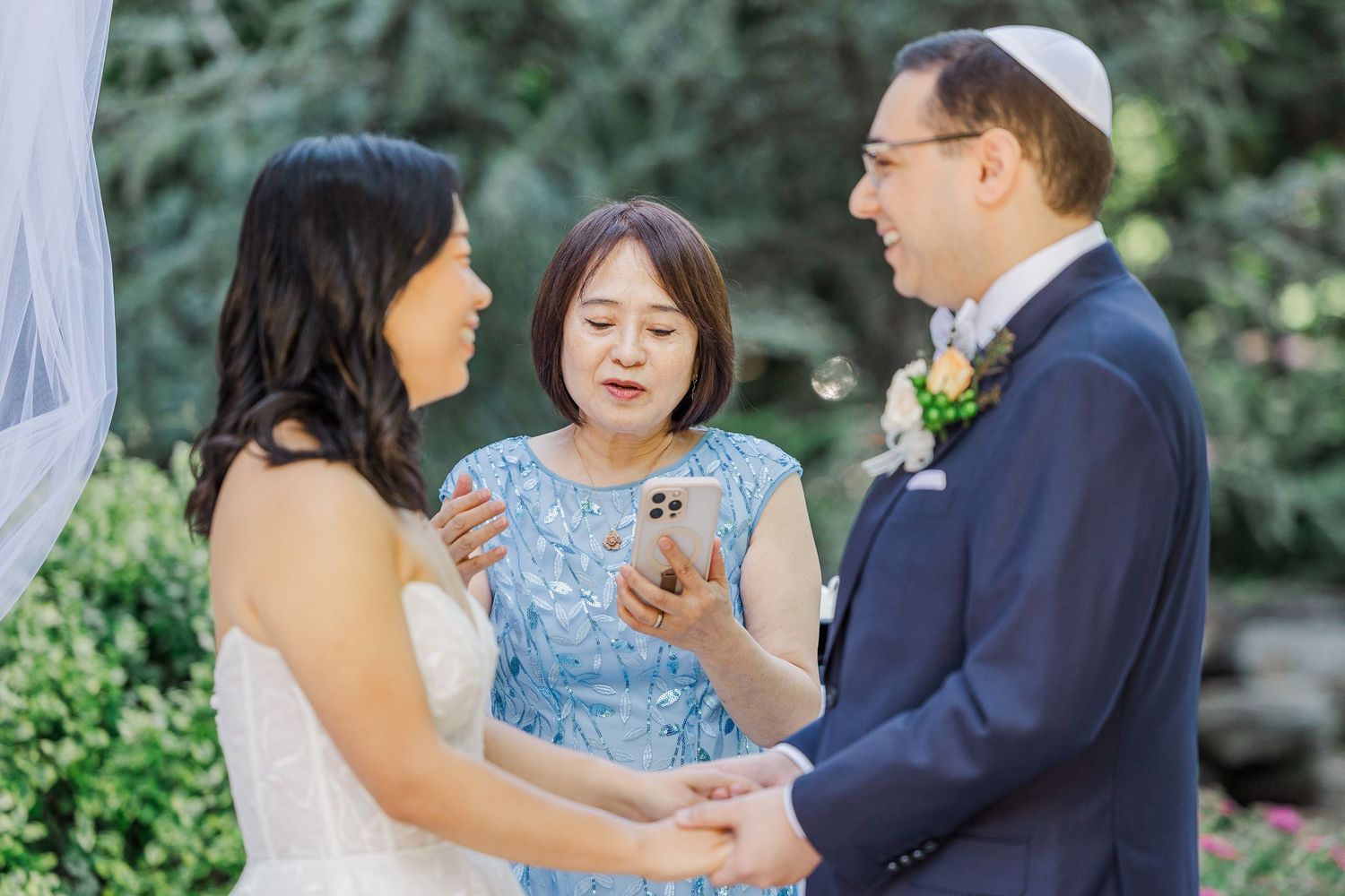Bride and groom exchange vows with officiant reading from phone at outdoor Jewish wedding ceremony.