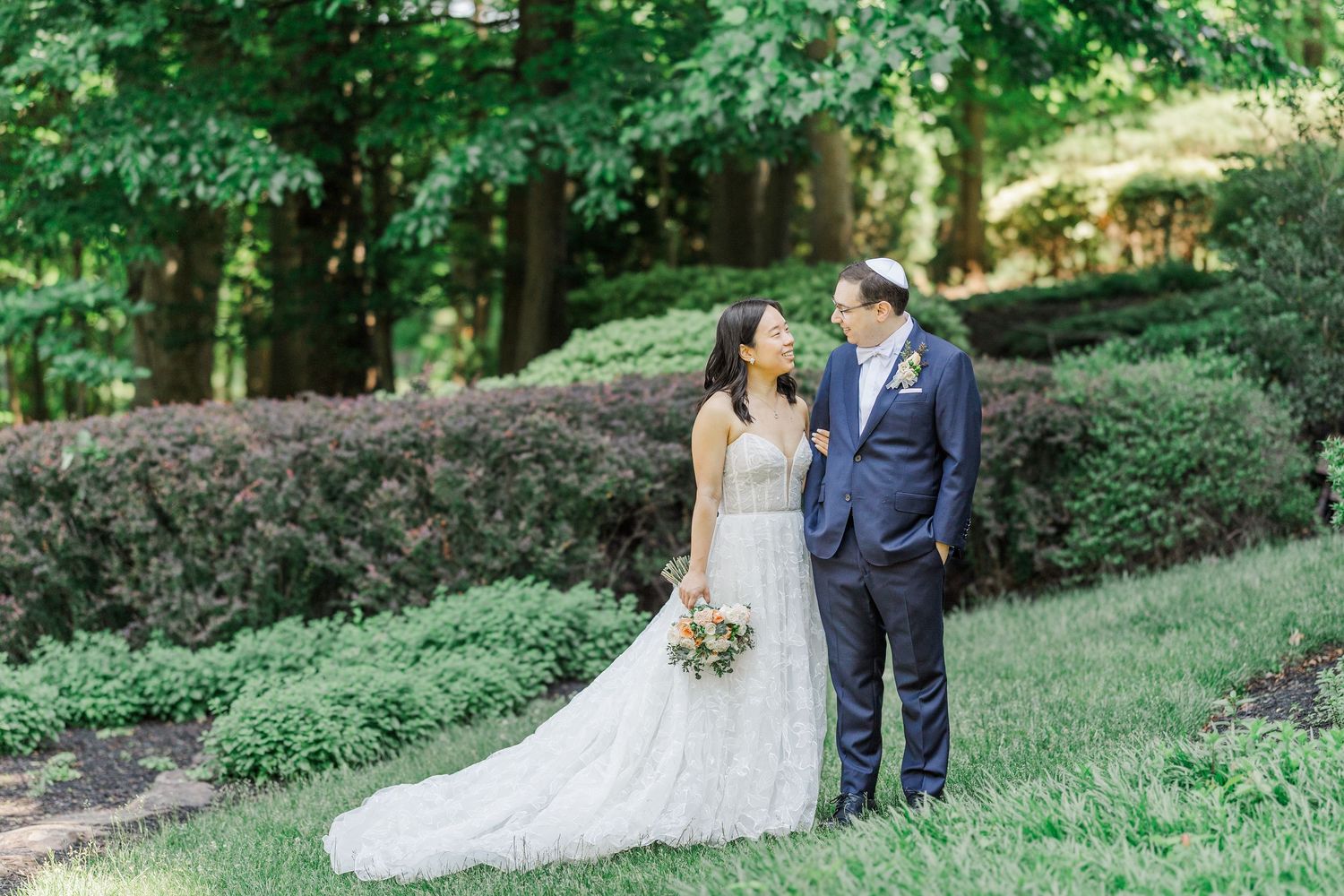 Bride and groom stand together on lush green lawn surrounded by manicured hedges and tall trees in an outdoor wedding portrait.