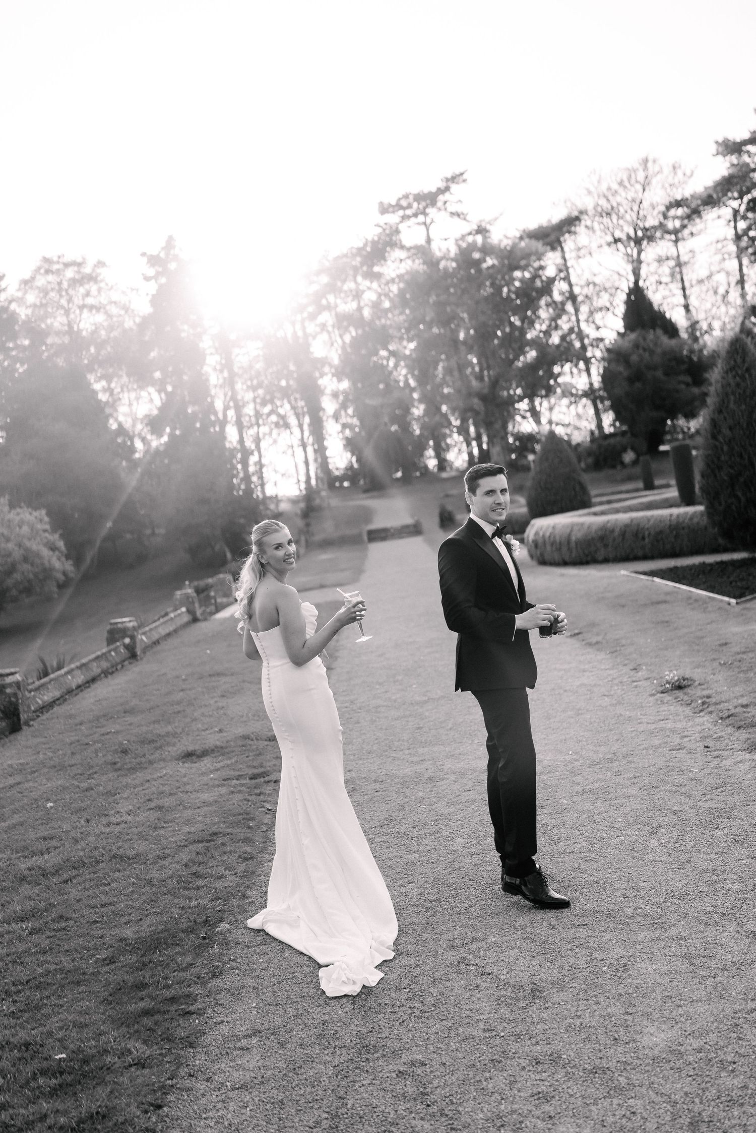 Black and white photo of a bride and groom holding champagne glasses outdoors at sunset in a garden.