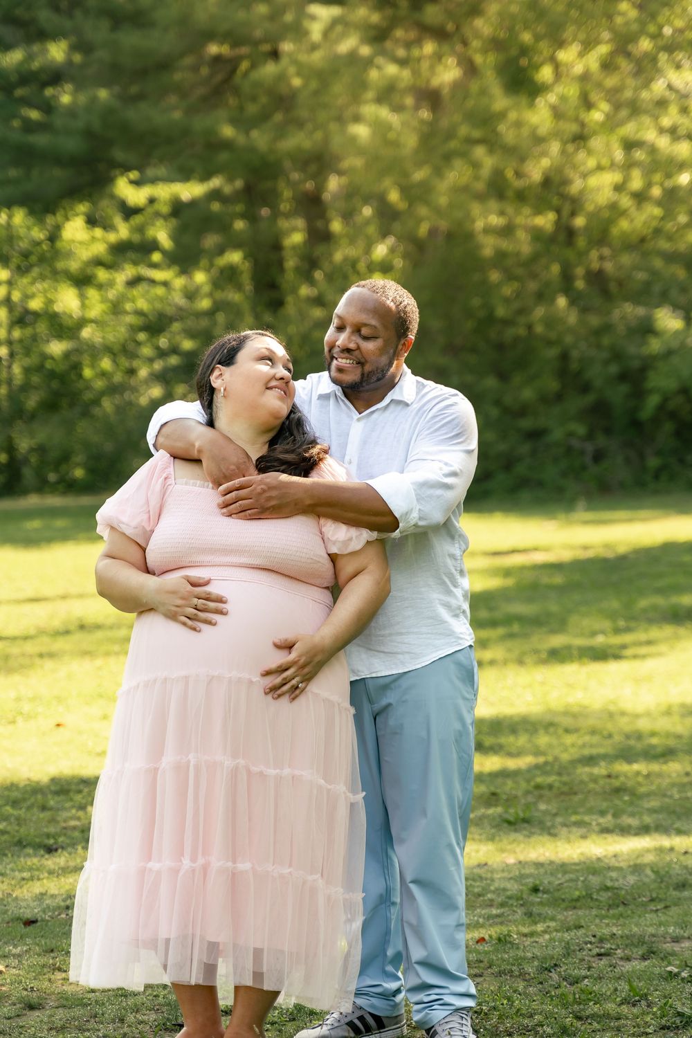 Maternity photo session, mom and dad  to-be embraced in the forest with sun shining behind them