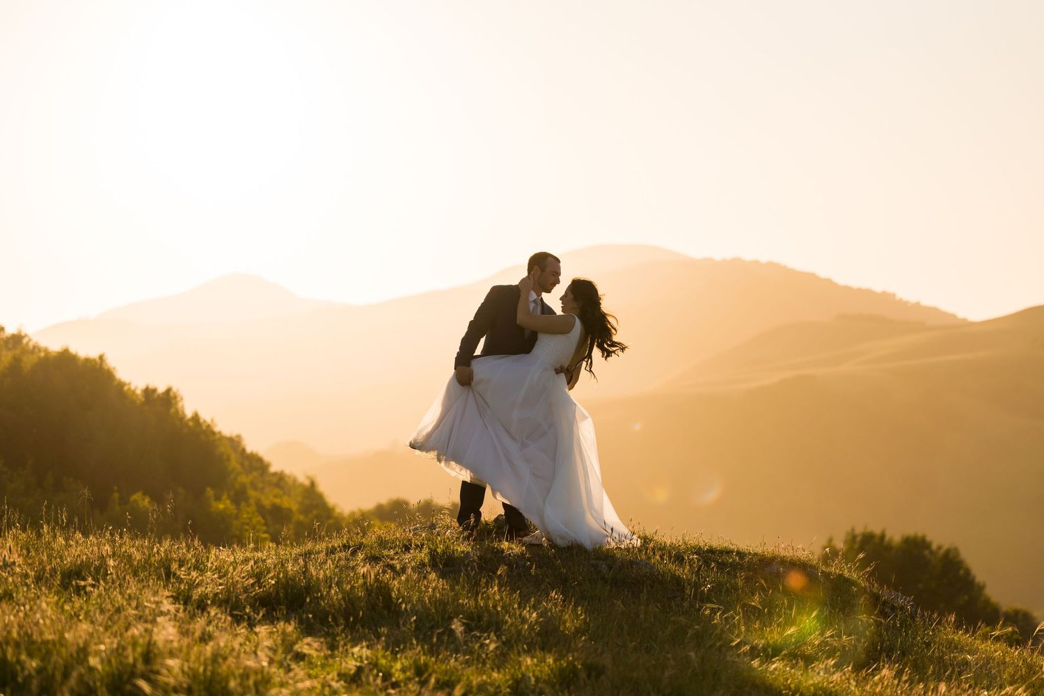 Couple sharing a romantic dip kiss on a sunlit mountain meadow at golden hour in wedding attire.
