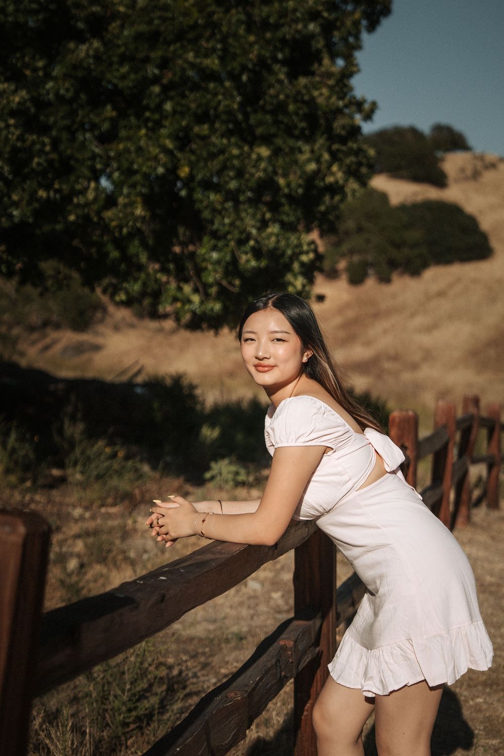 Young Asian woman in white dress leaning on wooden fence with golden hills and green trees in background.