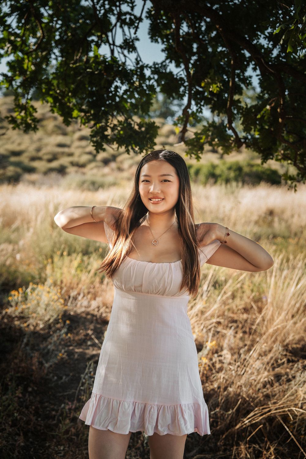 Young woman in white sundress smiling outdoors in golden field under tree during sunset.