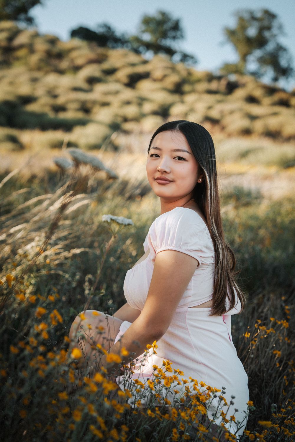 Young Asian woman in white dress sitting among yellow wildflowers in a sunlit field with rocky hills in the background.
