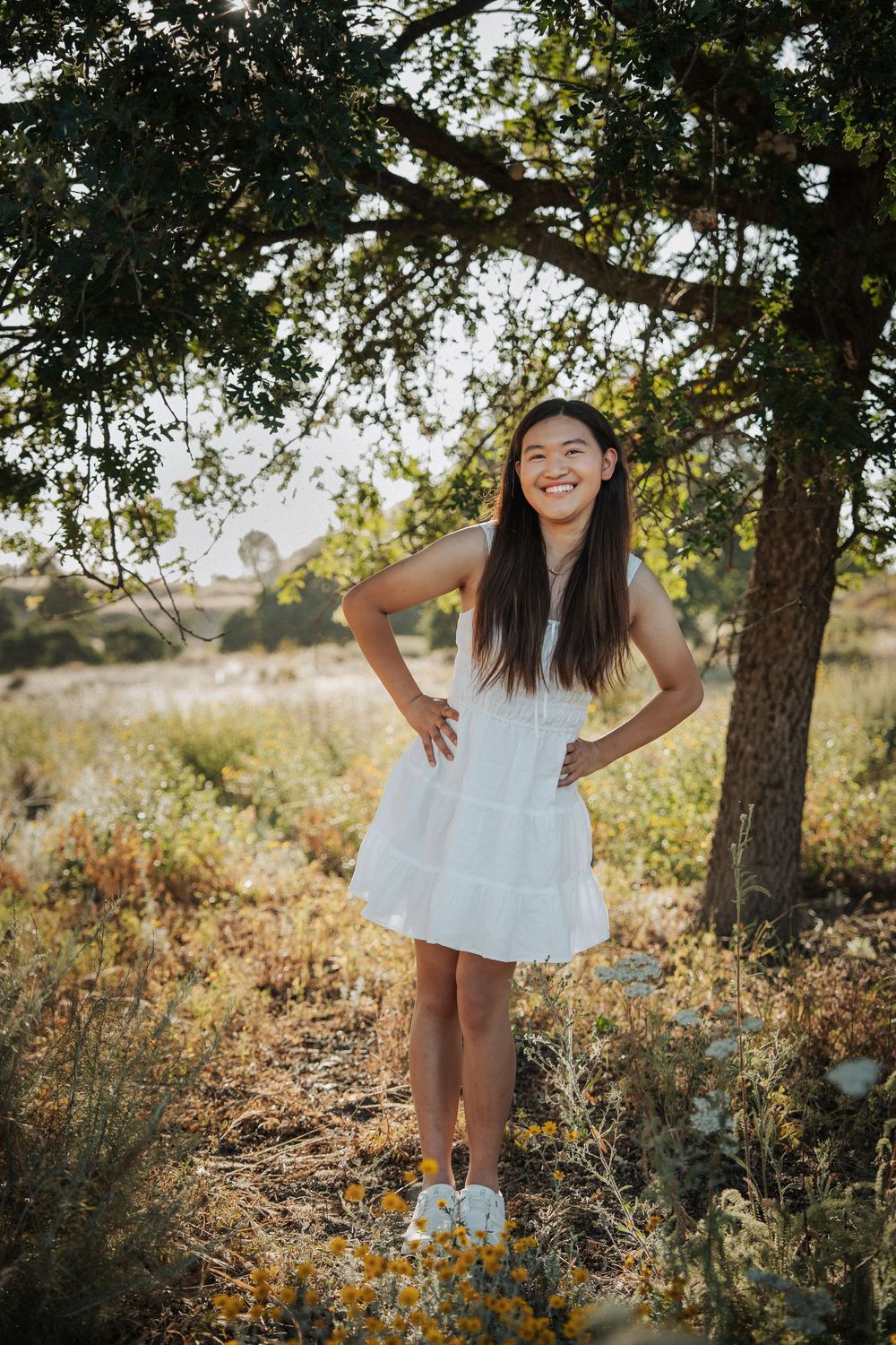Smiling young woman in white dress standing under a tree in a sunny field with hands on hips.