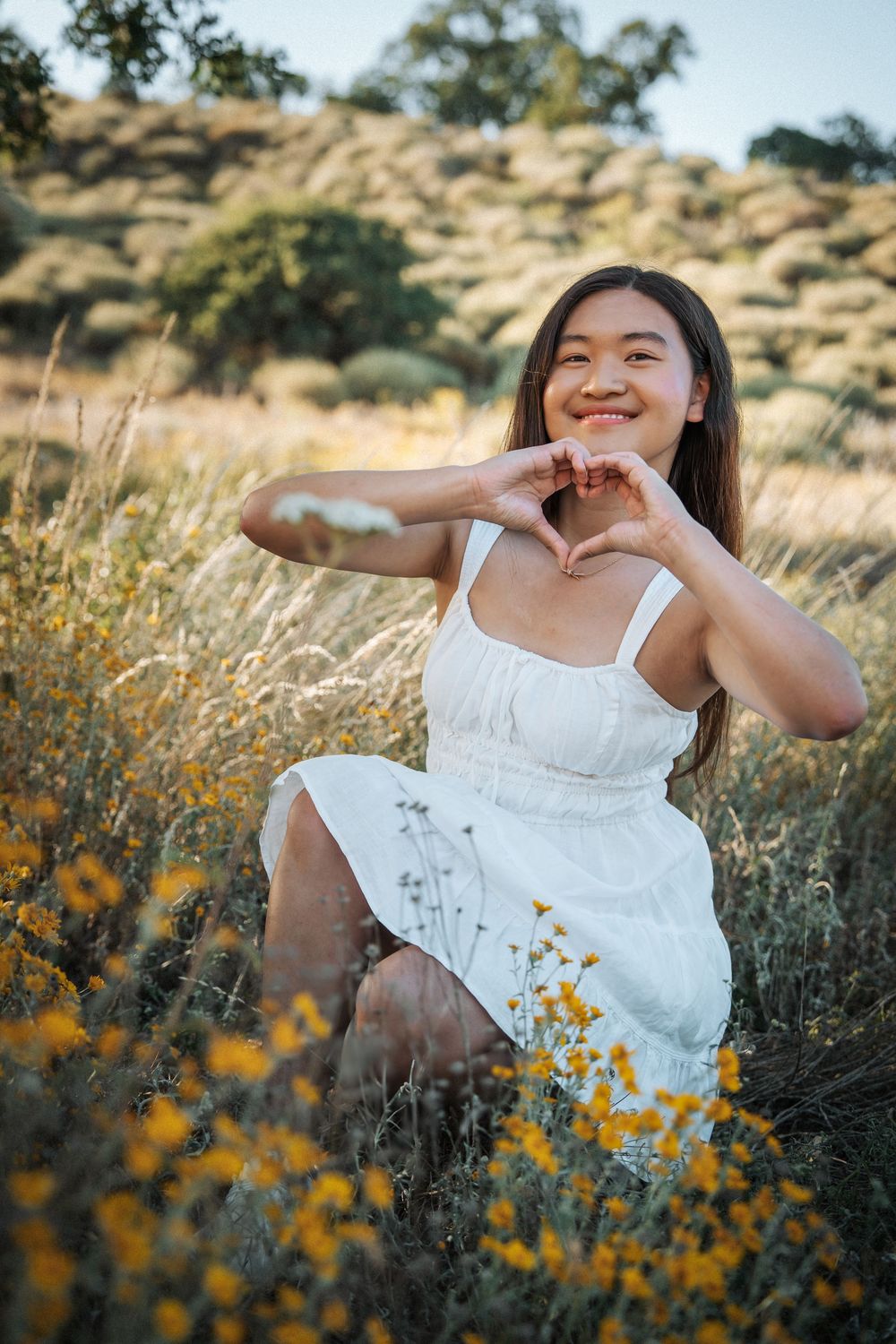 Young Asian woman in white dress sitting in golden field with yellow wildflowers, making a heart shape with her hands.